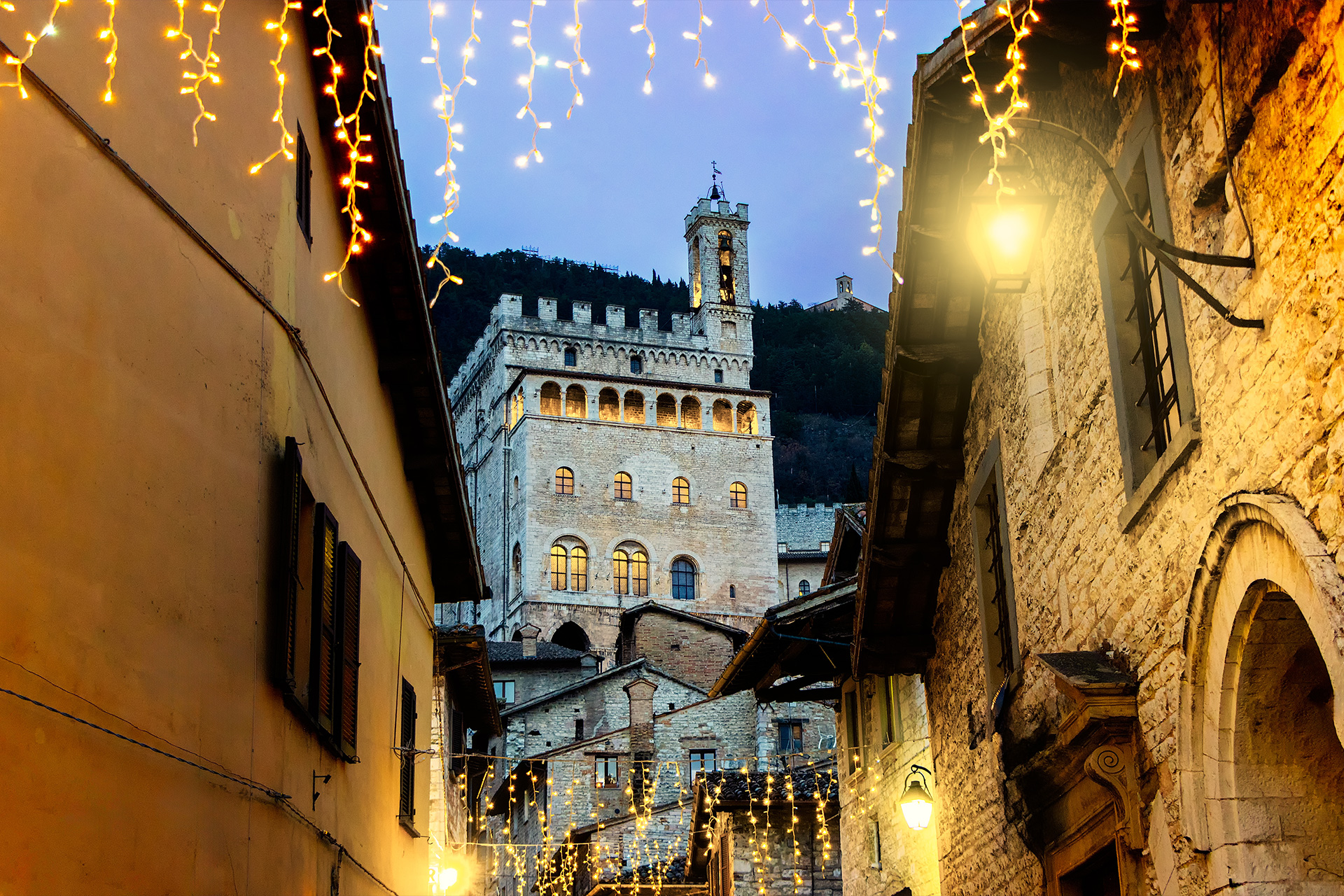 View of the Palazzo dei Consoli in Gubbio at dusk, framed by narrow stone streets and warm Christmas lights.