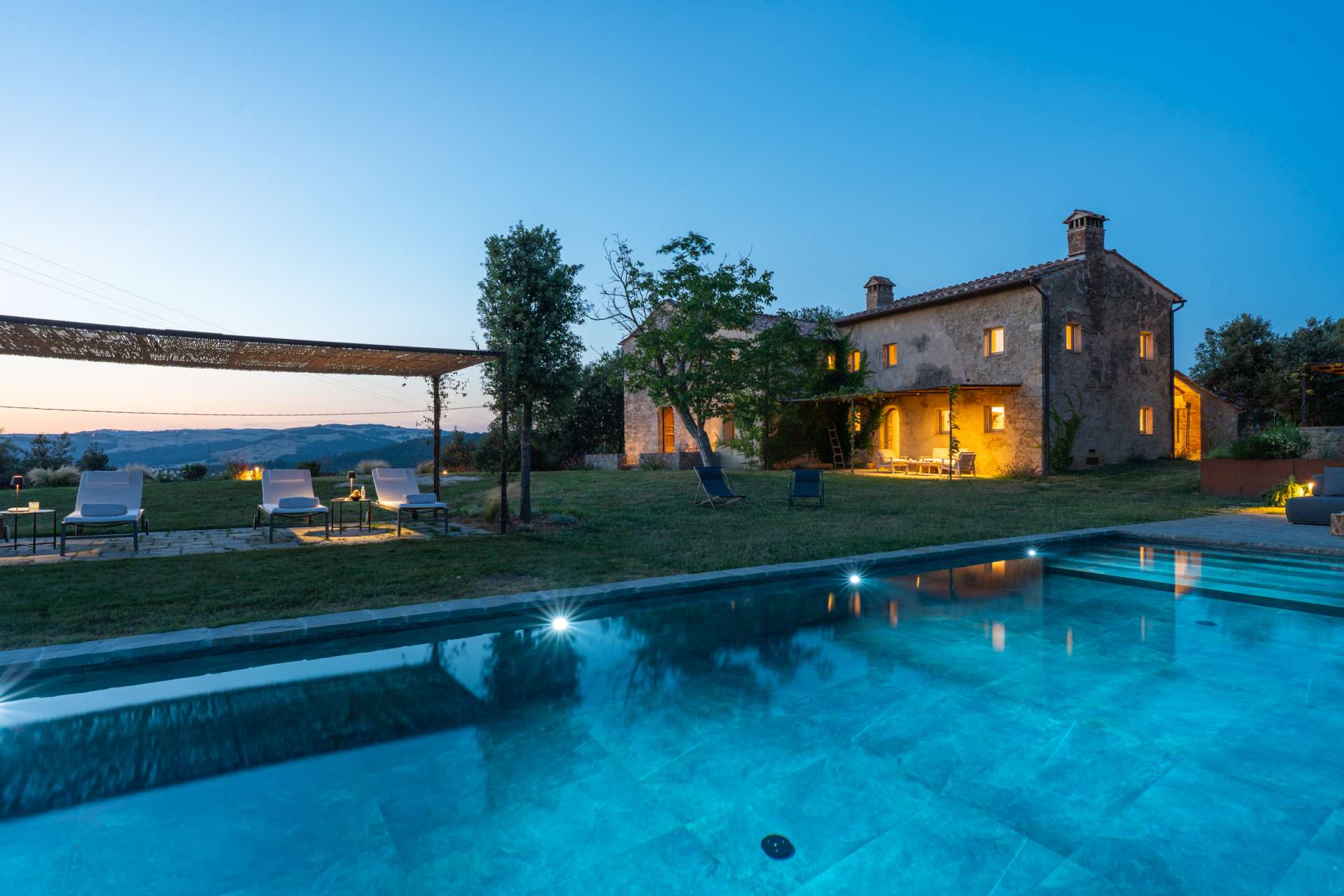 Evening view of Villa Pieve di Ponsano with illuminated stone farmhouse and private pool in Tuscany.