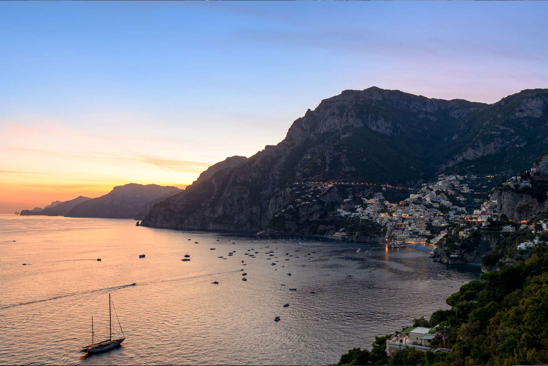 Sunset view over Positano with mountains, illuminated houses and boats on the Mediterranean Sea