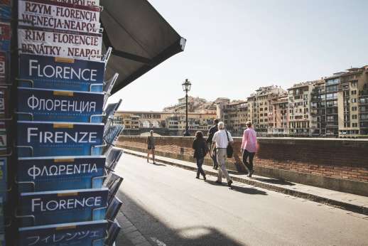 Arno Gondola Ride - A stack of tour guide books about Florence in multiple languages appear in a news stand while a tour group walk along the road by the Arno.