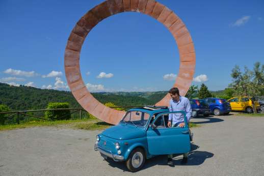 Fiat 500 Tour - A classic blue Fiat 500 with an open sunroof sits by a large, wheel-shaped monument overlooking the Chianti countryside