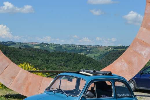 Fiat 500 Tour - A classic blue Fiat 500 with an open sunroof sits by a large, wheel-shaped monument overlooking the Chianti countryside
