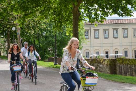 Lucca By Bike Or Foot - A group of people on rented bicycles cycle through a park in Lucca past an old building