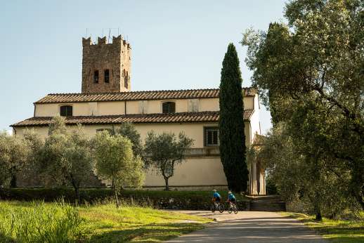 Two cyclists riding past a historic Tuscan church surrounded by olive trees and cypress trees in the countryside.